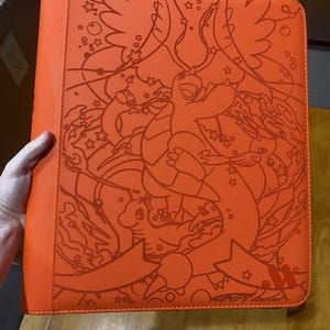 May include: A bright orange binder featuring a detailed line art illustration of a dragon-like creature with wings, surrounded by stars and swirling patterns. The cover has a textured surface and a ribbon-like graphic at the bottom right corner.