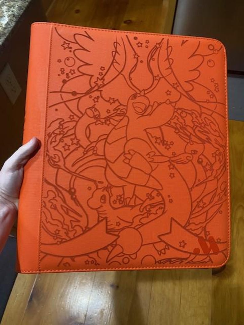 May include: A bright orange binder featuring a detailed line art illustration of a dragon-like creature with wings, surrounded by stars and swirling patterns. The cover has a textured surface and a ribbon-like graphic at the bottom right corner.
