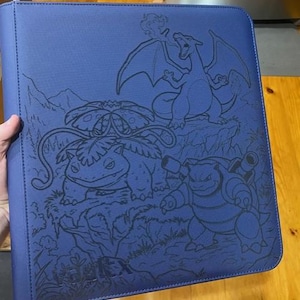 May include: A blue zipped binder featuring a dark blue embossed graphic of three creatures, including a winged dragon, a plant-based creature with a large flower, and a turtle-like creature with cannons, with the text Vault X at the bottom.