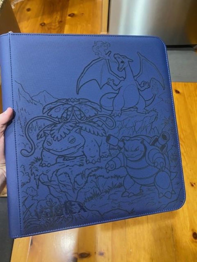 May include: A blue zipped binder featuring a dark blue embossed graphic of three creatures, including a winged dragon, a plant-based creature with a large flower, and a turtle-like creature with cannons, with the text Vault X at the bottom.