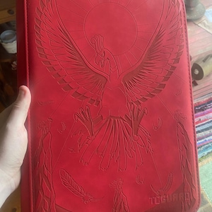 May include: A bright red rectangular trading card binder featuring a large embossed graphic of a phoenix-like bird with wings spread wide against a circular sun background. The word TCGUARD is embossed in the bottom right corner of the cover.