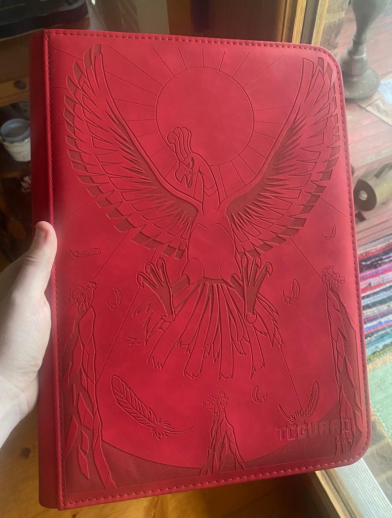 May include: A bright red rectangular trading card binder featuring a large embossed graphic of a phoenix-like bird with wings spread wide against a circular sun background. The word TCGUARD is embossed in the bottom right corner of the cover.