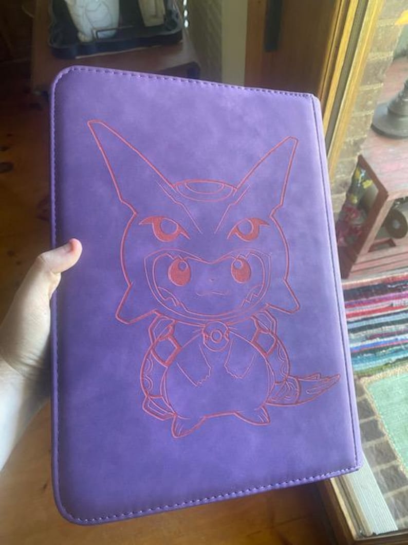 May include: A rectangular purple binder featuring a laser-engraved outline of a character wearing a hooded costume. The design depicts a small creature with pointed ears and a round body, centred on the front cover with visible stitching along the edges.