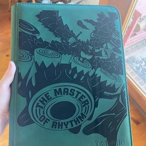 May include: A teal card binder featuring a black graphic of a primate playing drums with the text THE MASTER OF RHYTHM printed in a circular design. The bottom right corner displays the TCGUARD logo and the text guard your trading cards.