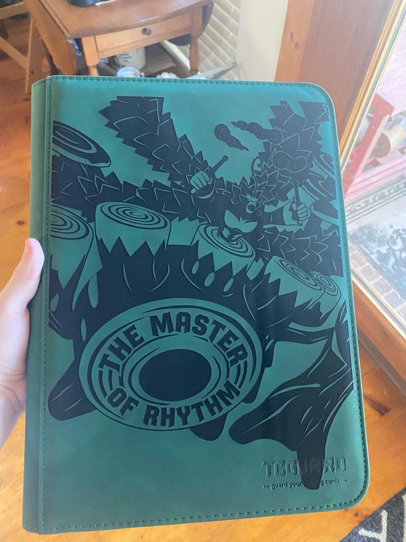 May include: A teal card binder featuring a black graphic of a primate playing drums with the text THE MASTER OF RHYTHM printed in a circular design. The bottom right corner displays the TCGUARD logo and the text guard your trading cards.