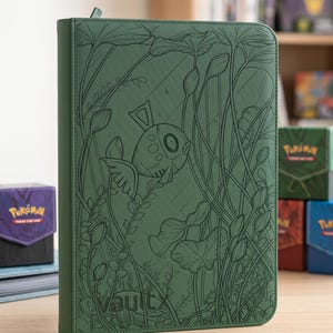 May include: A sage green zippered trading card binder featuring an embossed line art illustration of a fish swimming among aquatic plants and lily pads. The word vaultx is debossed in the bottom left corner of the textured synthetic cover.