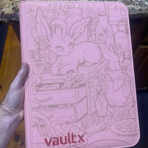 May include: A light pink zipped binder featuring a debossed illustration of a creature with large ears and a long tail surrounded by plants and bottles. The text vaultx is printed in a dark pink font at the bottom of the front cover.