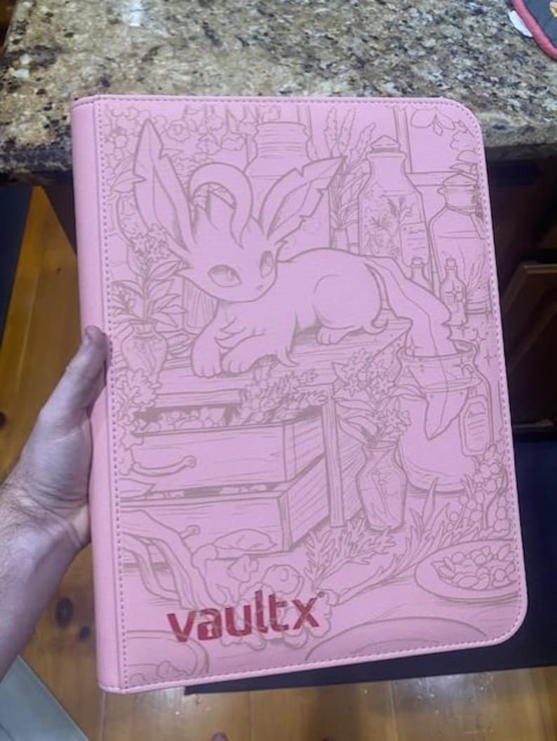 May include: A light pink zipped binder featuring a debossed illustration of a creature with large ears and a long tail surrounded by plants and bottles. The text vaultx is printed in a dark pink font at the bottom of the front cover.