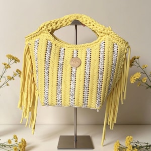 May include: A yellow handbag with a woven design and silver accents. The bag features a fringe detail and a wooden circular emblem. The handle is thick and braided. The bag is displayed on a metal stand.