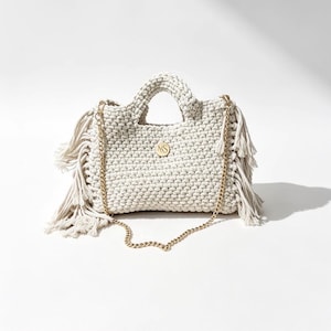 May include: A cream-coloured crocheted handbag featuring a textured knit pattern, two top handles, and long fringe detailing on both sides. The bag includes a gold-toned metal chain shoulder strap and a circular gold emblem with the letters MS.