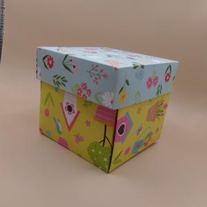 May include: A decorative box with a lid. The lid is light blue with a floral pattern of red, pink, and white flowers. The box is yellow with a pattern of birdhouses, butterflies, and gardening tools.