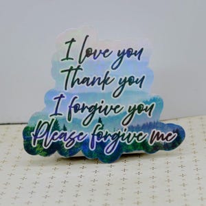 May include: Holographic sticker with the phrases "I love you", "Thank you", "I forgive you", and "Please forgive me" in black script. The sticker has a blue and green landscape design at the bottom.