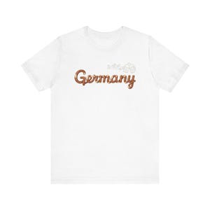 Op de afbeelding: Wit T-shirt met het woord "Germany" in een bruin, cursief lettertype. Boven het woord staat een kleine, lichtbruine illustratie van bierpullen en muzieknoten.