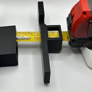 Puede incluir: Accesorio negro para herramientas de medición con una cinta métrica amarilla. La cinta métrica muestra 23 cm. El accesorio está diseñado para sujetar la cinta métrica en su lugar. La cinta métrica es roja y negra.