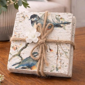 May include: A stack of square coasters with a bird and floral design, tied with twine and a white flower accent. The coasters feature a distressed, vintage look with blue, white, and brown tones. The coasters are on a wooden surface.