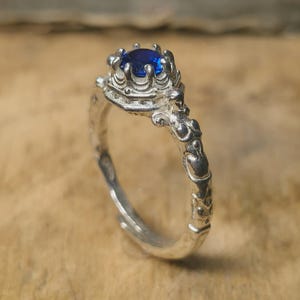 Op de afbeelding: Een zilveren ring met een centrale ronde blauwe edelsteen in een kroonvormige zetting. De ringband is voorzien van gedetailleerd, getextureerd metaalwerk met gebeeldhouwde figuren aan de zijkanten die naar de steenzetting toe leiden.