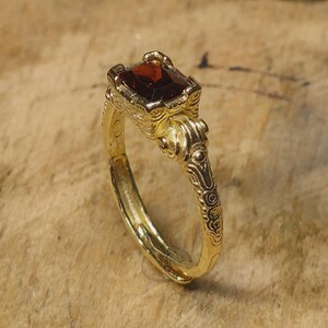 Puede incluir: Un anillo de oro con una piedra preciosa rectangular de color rojo intenso engastada en una montura elevada y ornamentada. La banda presenta intrincados patrones de volutas en relieve en los laterales, desde el engaste.
