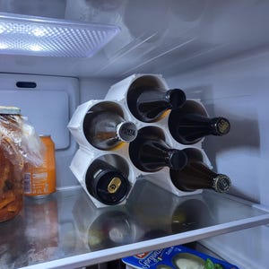 Può includere: Un portabottiglie esagonale bianco all'interno di un frigorifero. Il portabottiglie contiene sei bottiglie di vino. Una bottiglia ha un'etichetta "PINTA". Altri oggetti nel frigorifero includono un barattolo di cibo e una lattina arancione.