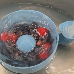 Puede incluir: Un dispositivo de lavado de verduras y frutas de color azul claro con una esfera central azul y una boquilla lateral. Se están lavando tomates rojos y productos oscuros con agua en el dispositivo, creando un movimiento arremolinado. El dispositivo está en un fregadero de acero inoxidable.