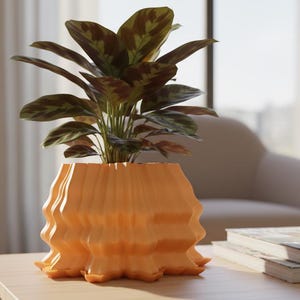 Peut inclure: Un pot de plante orange décoratif avec un design ondulé unique, contenant une plante à feuilles vertes et marron. Le pot est posé sur une table en bois clair, avec une pile de livres et une fenêtre en arrière-plan.