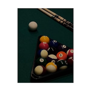 May include: A pool table scene featuring a triangle of numbered billiard balls in assorted colours, such as yellow, red, blue, and black. A white cue ball is positioned separately, with two pool cues above. The felt is green.