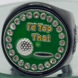 May include: Black ring with a green face featuring a golf ball design and the text "I'd Tap That" in yellow. The ring is encircled by a row of clear, sparkling stones.