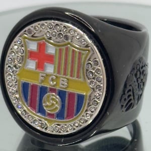 May include: A black ring featuring the FC Barcelona crest in the center. The crest is surrounded by small, clear stones. The ring has a glossy finish and a detailed design on the side.