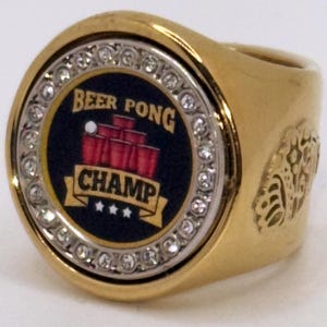 Puede incluir: Anillo dorado con una cara circular con las palabras "Beer Pong Champ" y un gráfico de vasos rojos y una pelota de ping pong. El anillo está adornado con pedrería transparente y tiene un diseño ornamentado en el lateral.