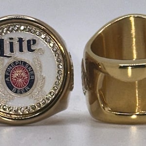 Peut inclure: Deux bagues dorées. L'une présente une face blanche avec le mot "Lite" en bleu et les mots "A Fine Pilsner Beer" dans un motif circulaire. L'autre bague est de couleur or massif avec un décapsuleur.
