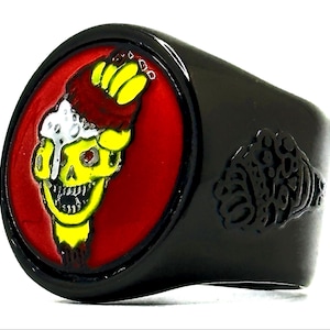 May include: Black ring with a skull design. The skull is yellow with a red eye, a red and brown hat, and a white substance dripping down. The skull is set against a red background. A black fist design is on the side of the ring.
