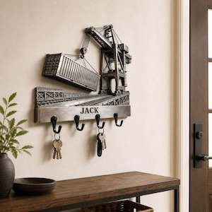 May include: A silver-coloured wall-mounted key holder depicting a crane lifting a shipping container. The key holder has four black hooks and the name "JACK" printed on it. Keys are hanging from three of the hooks.
