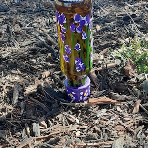 May include: A repurposed glass bottle, painted with purple irises and green stems, sits atop a small purple base. The bottle is brown and has a metallic cap. The base is decorated with white and yellow dots. The artwork is set on a bed of wood chips.