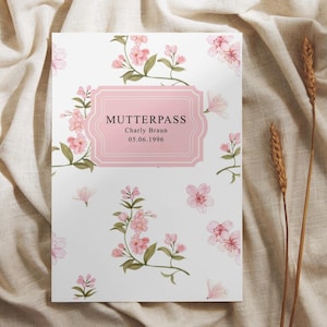 Könnte beinhalten: Ein rechteckiger Mutterpass-Umschlag mit weißem Hintergrund und einem wiederkehrenden Muster aus rosa Kirschblüten und grünen Blättern. Ein zentrales, rosafarbenes Etikett zeigt den Text MUTTERPASS, Charly Braun und 05.06.1996.