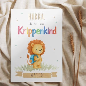Könnte beinhalten: Ein rechteckiger weißer Druck mit dem Text "HURRA. du bist ein Krippenkind" in bunten Buchstaben über einer Aquarellillustration eines Löwen mit blauem Rucksack. Der Name "MATEO" steht in Schwarz auf einem beigen Banner unten.