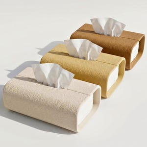 Puede incluir: Tres cajas de pañuelos rectangulares con superficie texturizada, dispuestas en fila diagonal. Las cajas son de color crema, amarillo mostaza y marrón, y cada una contiene una pila de pañuelos blancos que sobresalen por la ranura central.