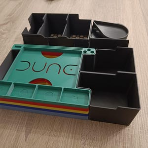 Könnte beinhalten: Ein Organizer für das Brettspiel Dune mit mehreren Fächern und einem teal-farbenen Tablett mit dem Namen des Spiels. Der Organizer ist hauptsächlich schwarz, mit bunten Schichten an der Basis. Er dient zur Aufbewahrung von Spielkomponenten.