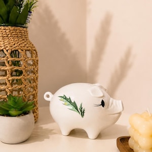 Può includere: Un salvadanaio in ceramica bianca con un disegno di foglia di agrifoglio verde dipinto sul lato. Il maialino ha una piccola coda circolare, orecchie a punta e occhi dipinti di nero con lunghe ciglia. Una statuetta liscia e lucida.