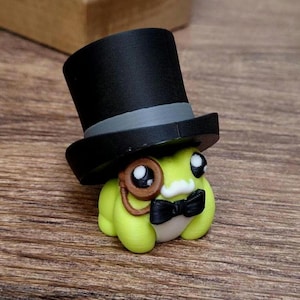 May include: A whimsical, light green frog figurine wearing a black top hat, a monocle, and a black bow tie. The frog has large, expressive eyes and is set against a wooden surface. A decorative wooden lantern is in the background.