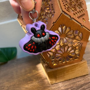 May include: A purple moth-shaped keychain with a silver clasp. The moth has black wings with red spots, a gray body, and red eyes. The keychain is held in front of a wooden lantern with intricate cut-out designs.