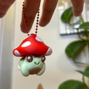May include: A whimsical keychain featuring a mushroom-frog hybrid. The red mushroom cap has white spots, and the frog is light green with a brown belly. The keychain is attached to a red beaded chain.