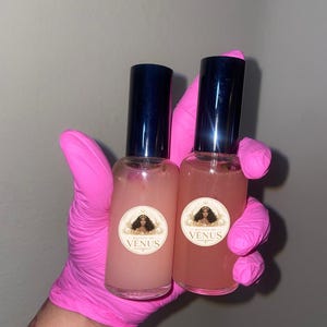 May include: Two clear glass spray bottles filled with pink liquid. Each bottle has a dark blue cap and a round label with a woman's portrait and the word "VENUS". The bottles are held by a person wearing pink gloves.