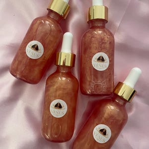 May include: Four bottles of shimmering body oil with white droppers and gold collars are arranged on a pink satin surface. Each bottle has a round label with the word "VENUS" and an illustration. The oil has a reddish-orange hue.