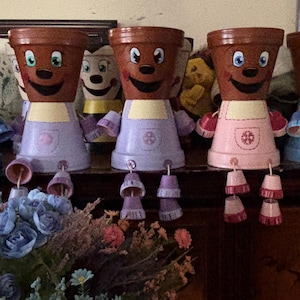 Może przedstawiać: Pięć fantazyjnych, antropomorficznych doniczek z uśmiechniętymi twarzami i kolorowymi fartuchami. Doniczki są brązowe z malowanymi cechami, a fartuchy są w odcieniach niebieskiego, różowego i fioletowego. Każda doniczka ma zwisające akcesoria w kształcie dzwonka. Bukiet kwiatów jest na pierwszym planie.