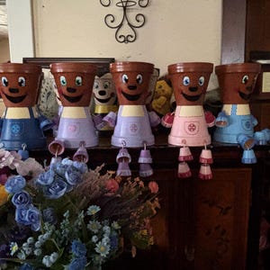 May include: Five whimsical, anthropomorphic flower pots with smiling faces and colorful aprons. The pots are brown with painted features, and the aprons are blue, lavender, and pink. Each pot has dangling bell-shaped accessories. A bouquet of colorful flowers is in the foreground.