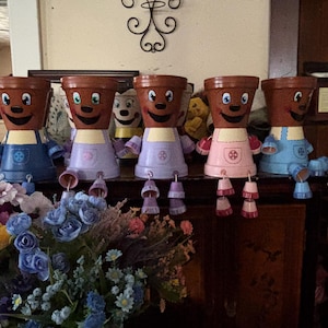 Puede incluir: Cinco macetas antropomórficas caprichosas con caras sonrientes y delantales coloridos. Las macetas son marrones con rasgos pintados, y los delantales son en tonos de azul, rosa y morado. Cada maceta tiene accesorios colgantes en forma de campana. Un ramo de flores está en primer plano.