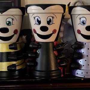 Peut inclure: Trois pots de fleurs peints à la main avec des motifs fantaisistes d'abeille et de vampire. Le pot d'abeille a des rayures jaunes et noires, le pot de vampire est noir et le troisième pot est violet avec des pois blancs. Chaque pot présente un visage souriant et des accents décoratifs.