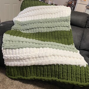 May include: A chunky knit blanket in shades of green and white. The blanket features thick, textured yarn in stripes and diagonal patterns. The color palette includes a deep forest green, a lighter sage green, and a bright white.