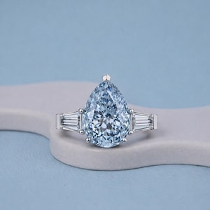 以下が含まれることがあります： 大きなペアシェイプのライトブルーダイヤモンドが特徴の、見事な婚約指輪。 リングはプラチナバンドで、メインストーンの両側に長方形のバゲットダイヤモンドが2つ付いています。 リングは水色の背景にセットされています。