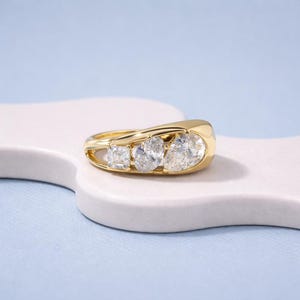 Könnte beinhalten: Ein goldener Ring mit drei klaren Edelsteinen. Der Ring hat ein glattes, poliertes Band und ein modernes Design. Die Edelsteine sind in einer Reihe angeordnet, mit einem quadratisch geschliffenen Stein links und zwei oval geschliffenen Steinen in der Mitte.