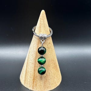 May include: A silver keychain with three green and black tiger eye beads. The beads are round and hang from a decorative silver loop. The keychain is displayed on a wooden cone against a black background.